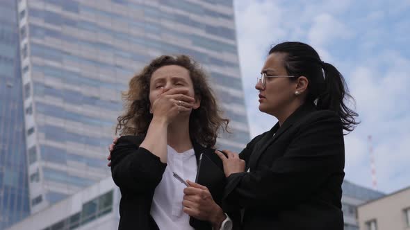 Brunette Corporate Worker Cheers Up Her Stressed Blonde Colleague in Front of Skyscraper