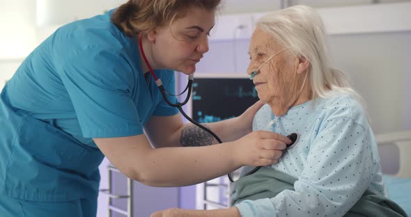 Old Woman Lying in Bed in Hospital Ward Being Examined By Doctor Using Stethoscope