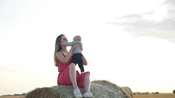 View of Happy Mother Throwing Up and Kissing Her Baby Son on Haystack at Sunset