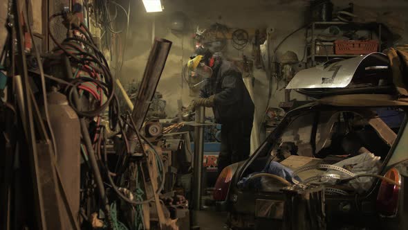 Man angel grinds metal plate on steel piping in messy workshop. WIDE SHOT