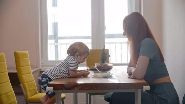 Ginger Woman Mother Sits By the Table with Her Little Baby Who Tries to Steal Apple Pieces From the