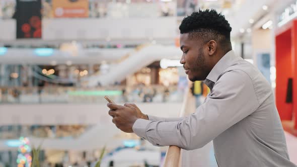 Young African American Man Standing in Shopping Mall Spending Free Time on Social Media Internet on