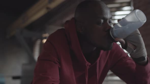 Tired African Boxer in Hoodie Leaning on Ropes and Drinking Water