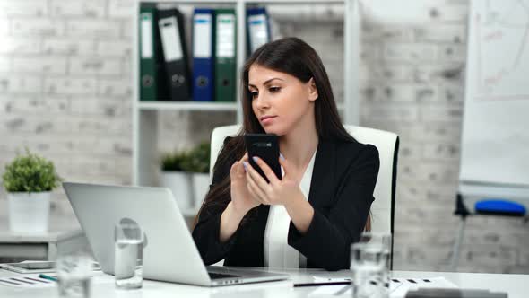 Confident Business Female Typing Number Calling Using Smartphone Looking at Screen of Laptop