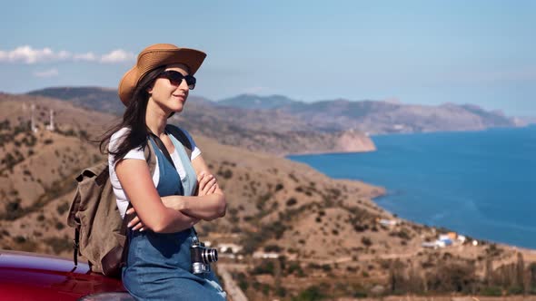 Beautiful Young Female Tourist Sitting on Car Bonnet with Crossed Hands Relaxing at Summer Seascape