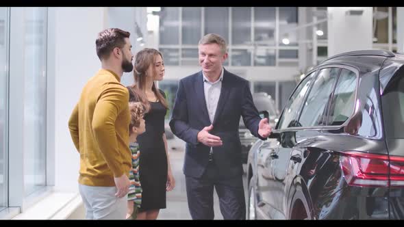 Confident Male Car Dealer Advertising New Car To the Caucasian Family in Automobile Showroom and