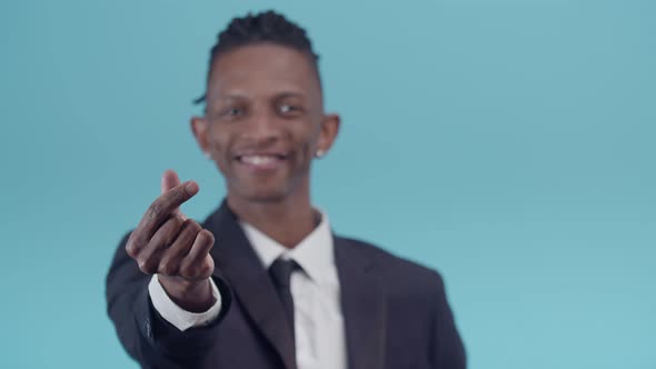 Black Man in an Office Suit Smiling Makes a Gesture of Money with Fingers on a Blue Studio