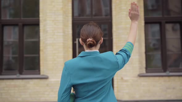 Back View of Slim Brunette Stewardess Standing in Front of Building and Waving. Young Caucasian