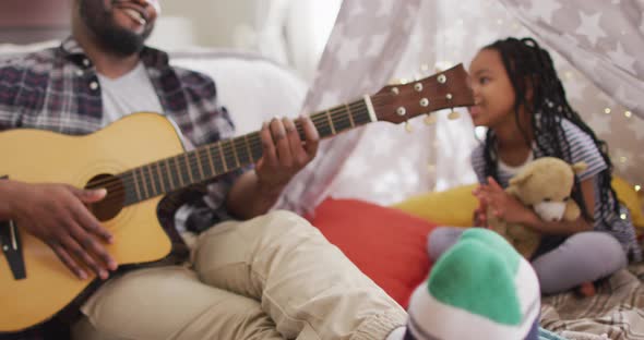 Happy african american father and daughter sitting in makeshift tent, playing guitar and singing