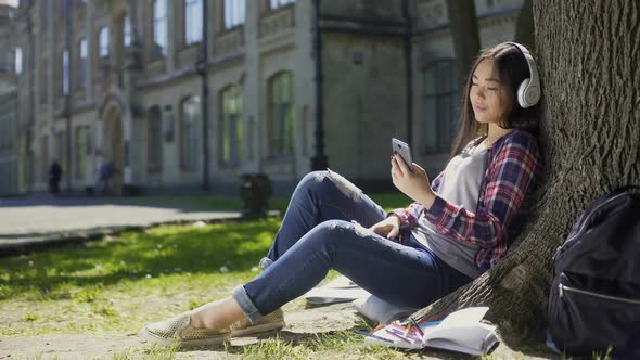 Mixed-Race Girl Sitting Under Tree in Headphones, Scrolling Screen of Cellphone