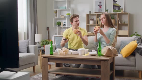 Young Couple Eating a Hamburger and Watching TV in the Living Room