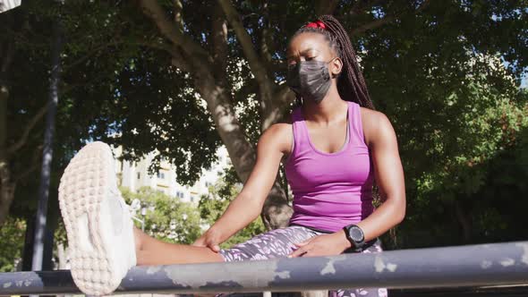 African american woman wearing face mask stretching her leg on the railing outdoors