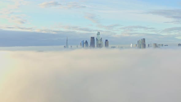 Dolly back aerial shot over morning fog towards London city centre skyscrapers sunrise