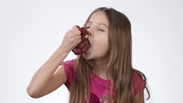 Little Girl Eating Ripe Big Grapes on a White Background. The Girl Appetizingly Examines the Grapes