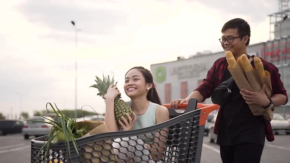 Likable Young Asian Woman Sitting in the Shopping Trolley with Paper Bags