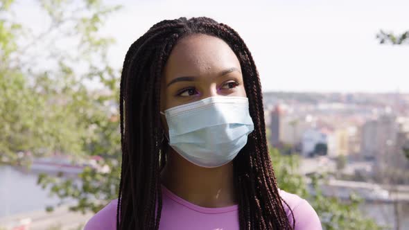 A Young Black Woman in a Face Mask Thinks About Something - Face Closeup - Trees and a Cityscape