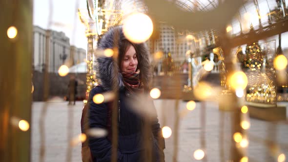 Portrait of a beautiful young woman in a jacket and hood through a garland on the street.