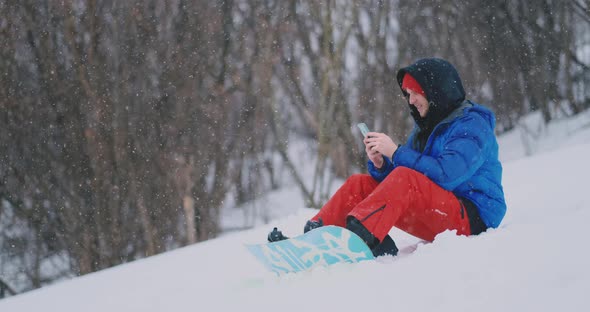 A Male Snowboarder Sitting on the Snow Takes Photos on the Phone of a Beautiful Resort Landscape