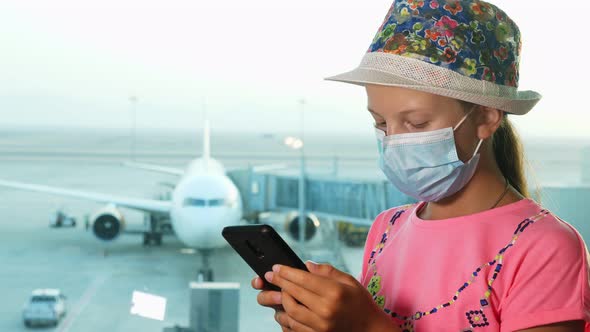 Teenage Girl, in Protective Mask, Uses Mobile, in Front of Panoramic Window with Runway 