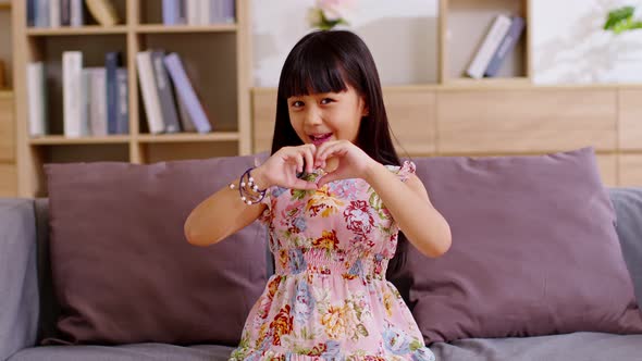 Asian kid sitting in couch and showing heart gesture with fingers image. Little girl smiling posing