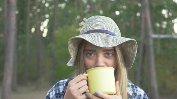 Young Happy Caucasian Girl in Plaid Shirt and Hat Drinking Tea or Coffe From the Yellow Cup, Smiling