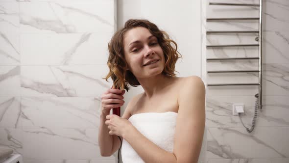 Smiling Young Woman in Big White Bath Towel Styling Her Hair with a Curling Iron in Front of a