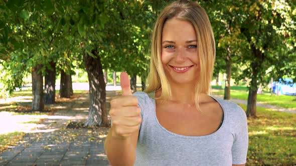 Young Pretty Blond Woman Shows Thumb Up on Agreement - Park with Trees in Background