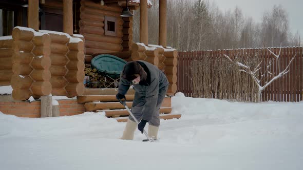 Man Cleans Shovel In Winter Yard From Snow On The Background Of A Wooden House