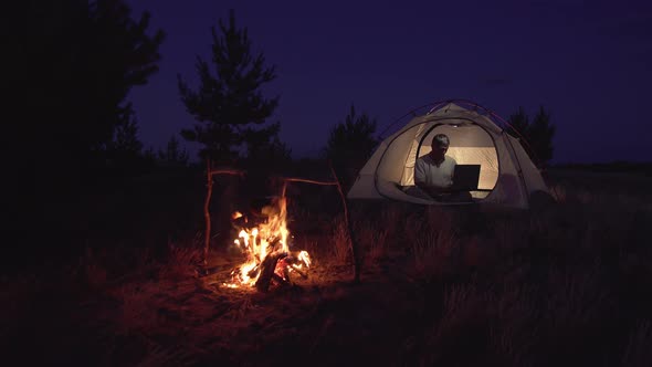 Freelancer Works Behind a Laptopin in a Tent Near a Fire