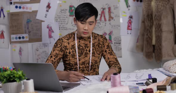 Asian clothing designer working on laptop in his office, creating a new clothes collection