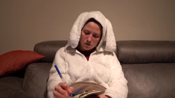 Woman dressed in rabbit pajamas writing in agenda with mobile on the sofa