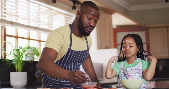 Happy african american father and daughter baking, preparing pizza in kitchen, adding tomato sauce