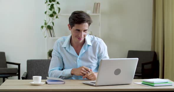 Young Attractive Smiling Man Sits at Table in Front of Laptop, Communicates Online with Webcam in