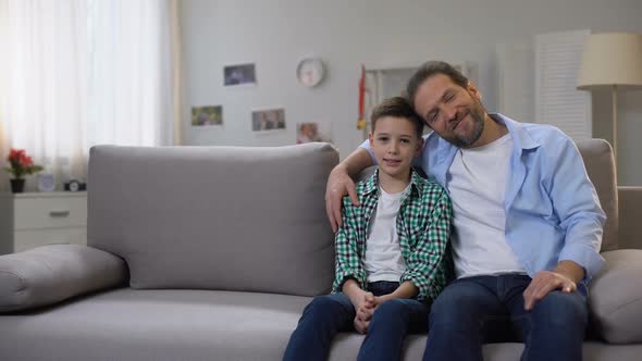 Happy Smiling Dad and Schoolboy Son Sitting on Sofa, Looking to Camera, Family