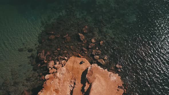 Young man with motorcycle helmet rests by the transparent sea, aerial view moving away from him