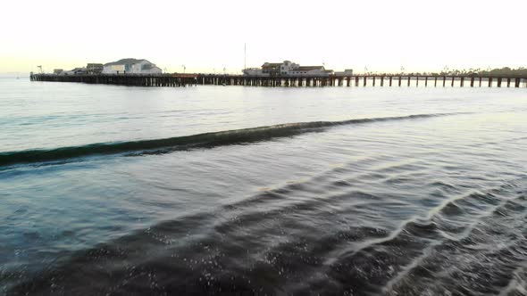 Aerial drone shot flying low over the waves and beach looking out at Stearns Wharf pier at sunset in