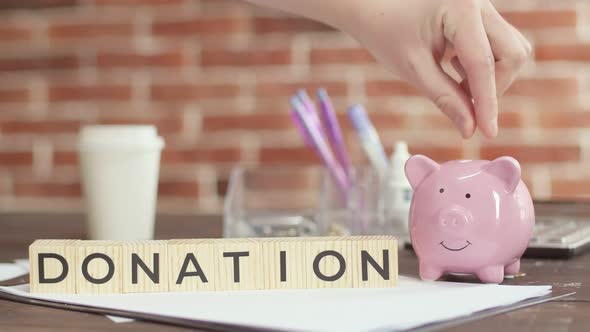 Donation is word written on wooden cubes lying on desk. Hand puts money in piggy bank.