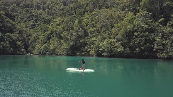 Aerial Shot: Woman on Paddleboard Feeling the Fresh Air in a Lagoon with Forest.