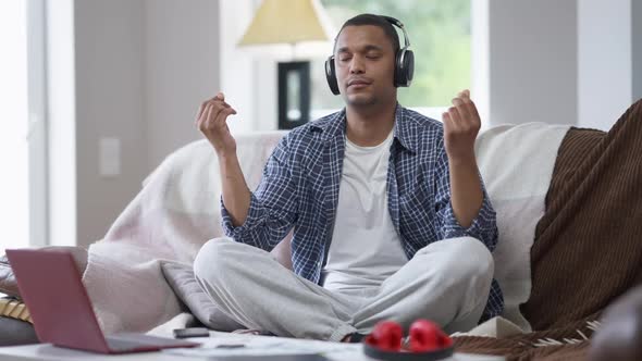 African American Man in Headphones Sitting on Couch in Lotus Pose Having Break in Home Office