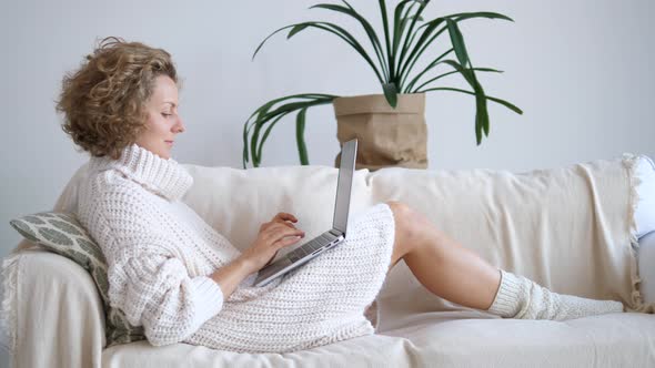 Young Woman In Knitted Sweater Using Laptop At Home Relaxing On Sofa