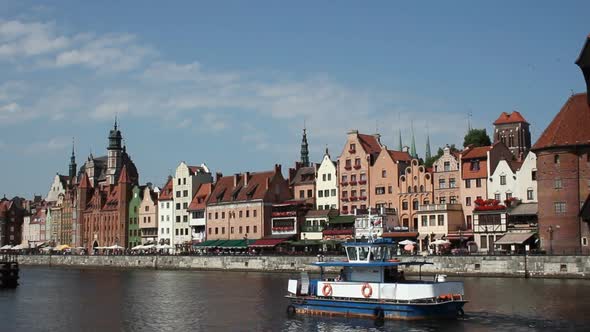 Small Ship Sailing by Riverbank with Row of Houses Aligned Along It, Polish City