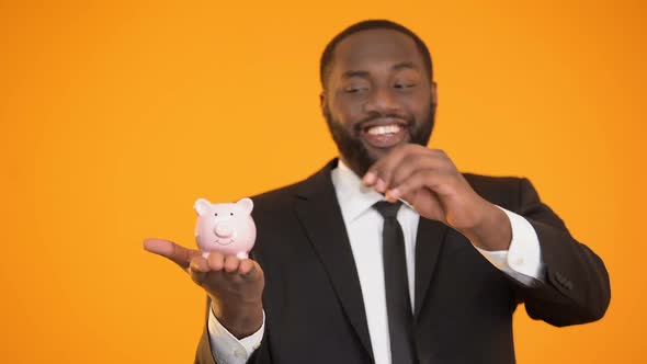 Smiling Black Man in Suit Putting Coin in Piggybank, Financial Literacy, Savings