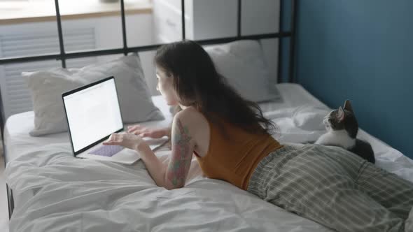 Young Asian Woman Working on Laptop Online at Home Lying on Bed in Bedroom with Her Cat
