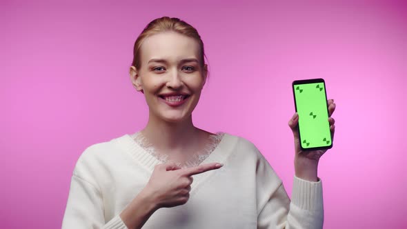 Young Attractive Woman Smiles While Pointing Finger at Smartphone with Green Screen Pink Background