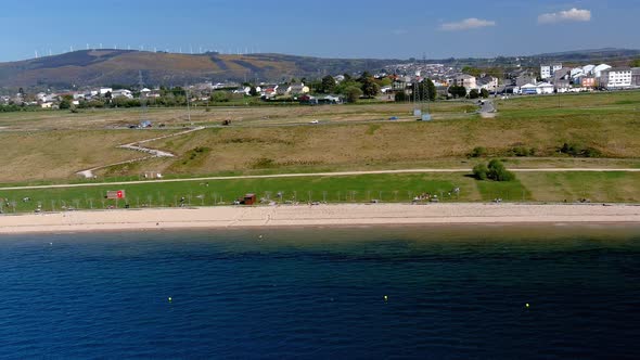 Lake beach with people sunbathing and bathing, garden and walking area, mountains in the background