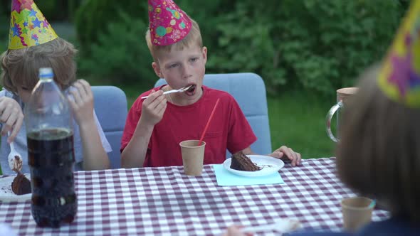 Redhead Caucasian Boy in Party Hat Eating Delicious Chocolate Birthday Cake Sitting with Friends on