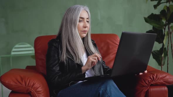 Stylish Woman with Gray Hair Sitting in Soft Chair and Having Video Chat on Computer