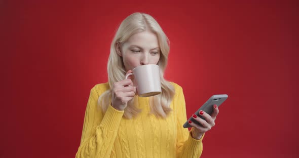 Beautiful Young Woman Drinks Cup Tea and Uses Phone Reads News Looks at the Camera in Surprise