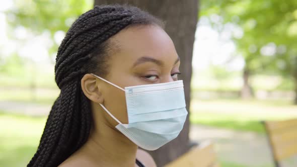 A Young Black Woman Puts on a Face Mask and Looks at the Camera As She Sits on a Bench