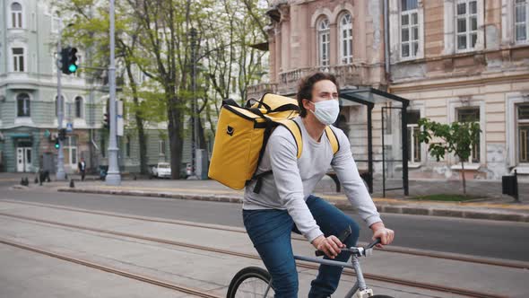 Poartrait of Courier with Yellow Backpack and Protective Mask Rides a Bicycle on the Street Through